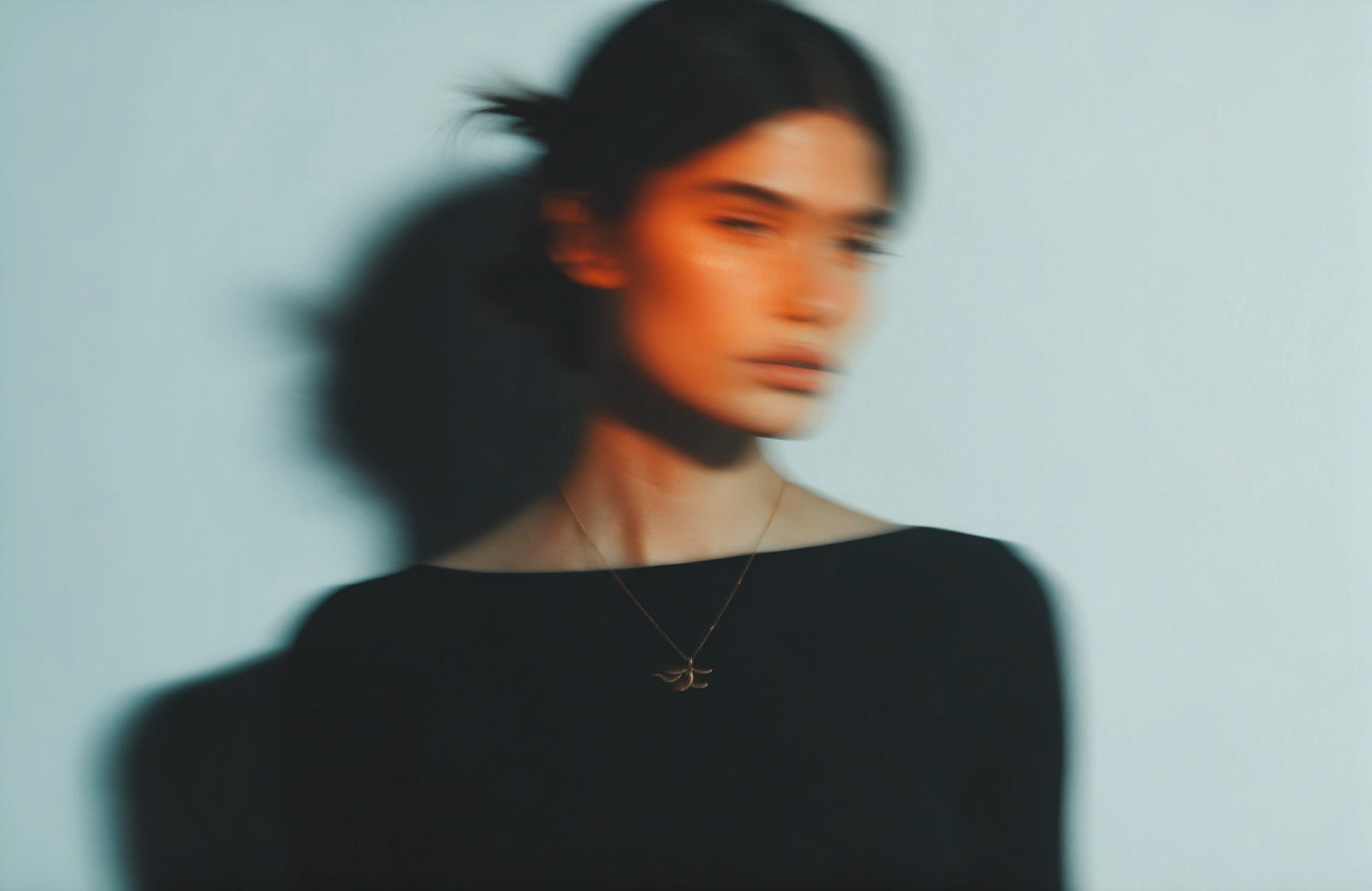A gold pendant necklace worn with a black top, lit warm against a pale wall